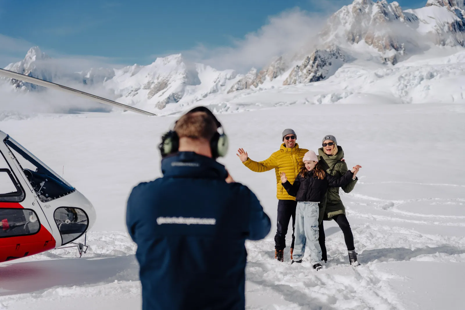 Guide in headset photographing three smiling visitors on snow beside a red and white helicopter