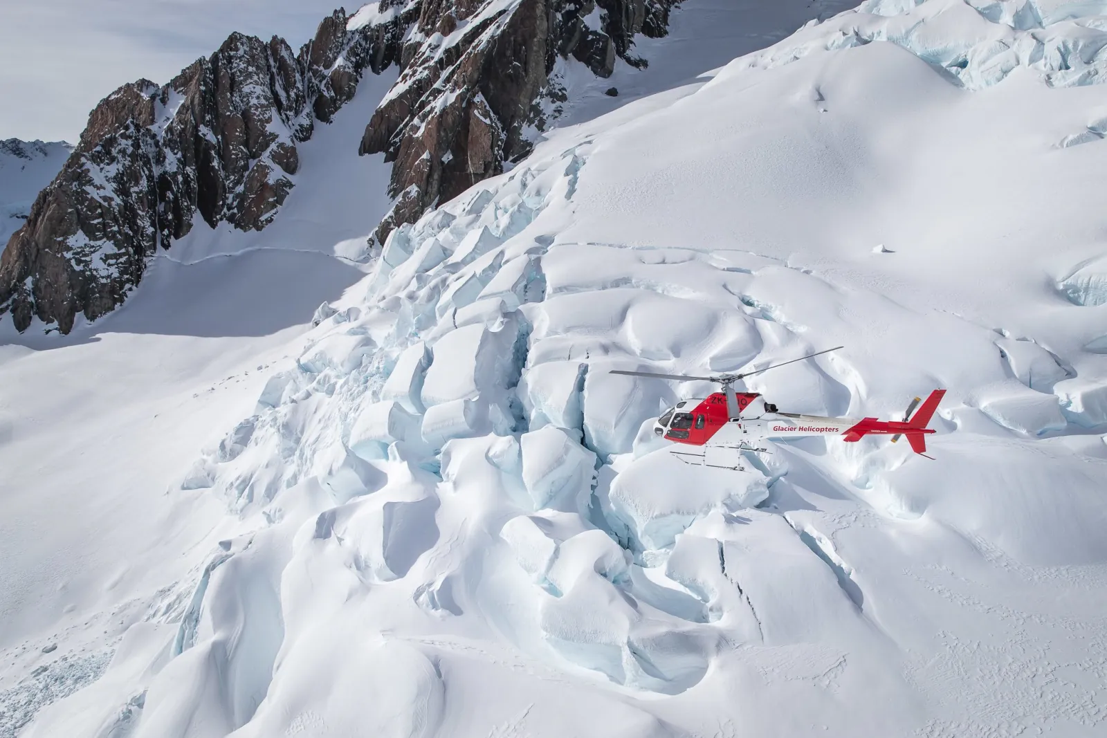 Red helicopter hovering over deep crevasses and wind-carved snow with rocky ridges above
