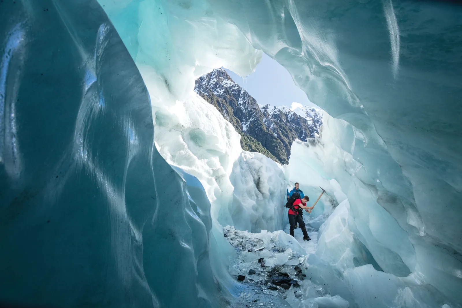 Lone hiker inside a large blue ice cave with an opening onto a rocky peak