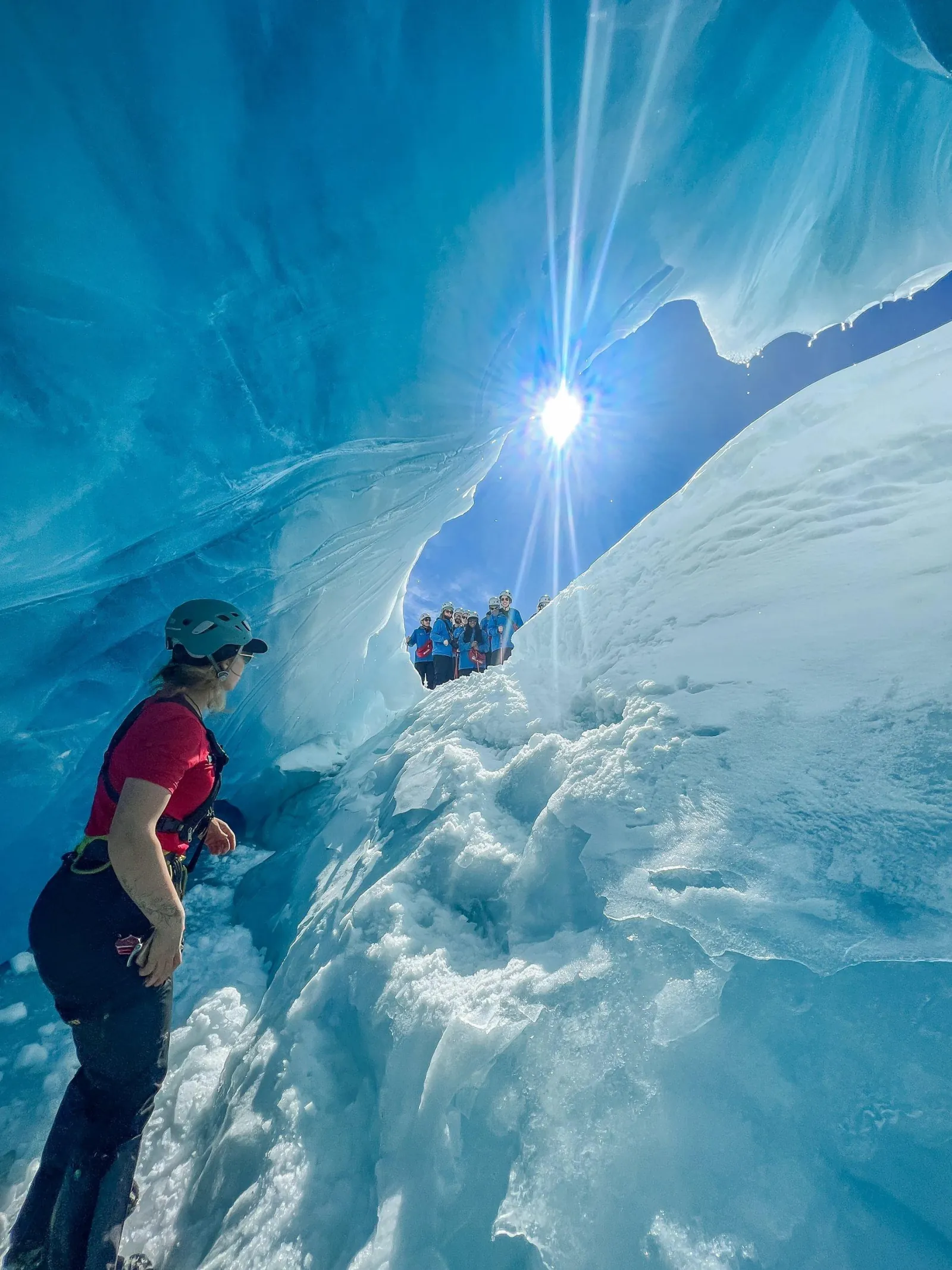 Hiker in red top looking through a blue ice gap with sunburst and snow slope