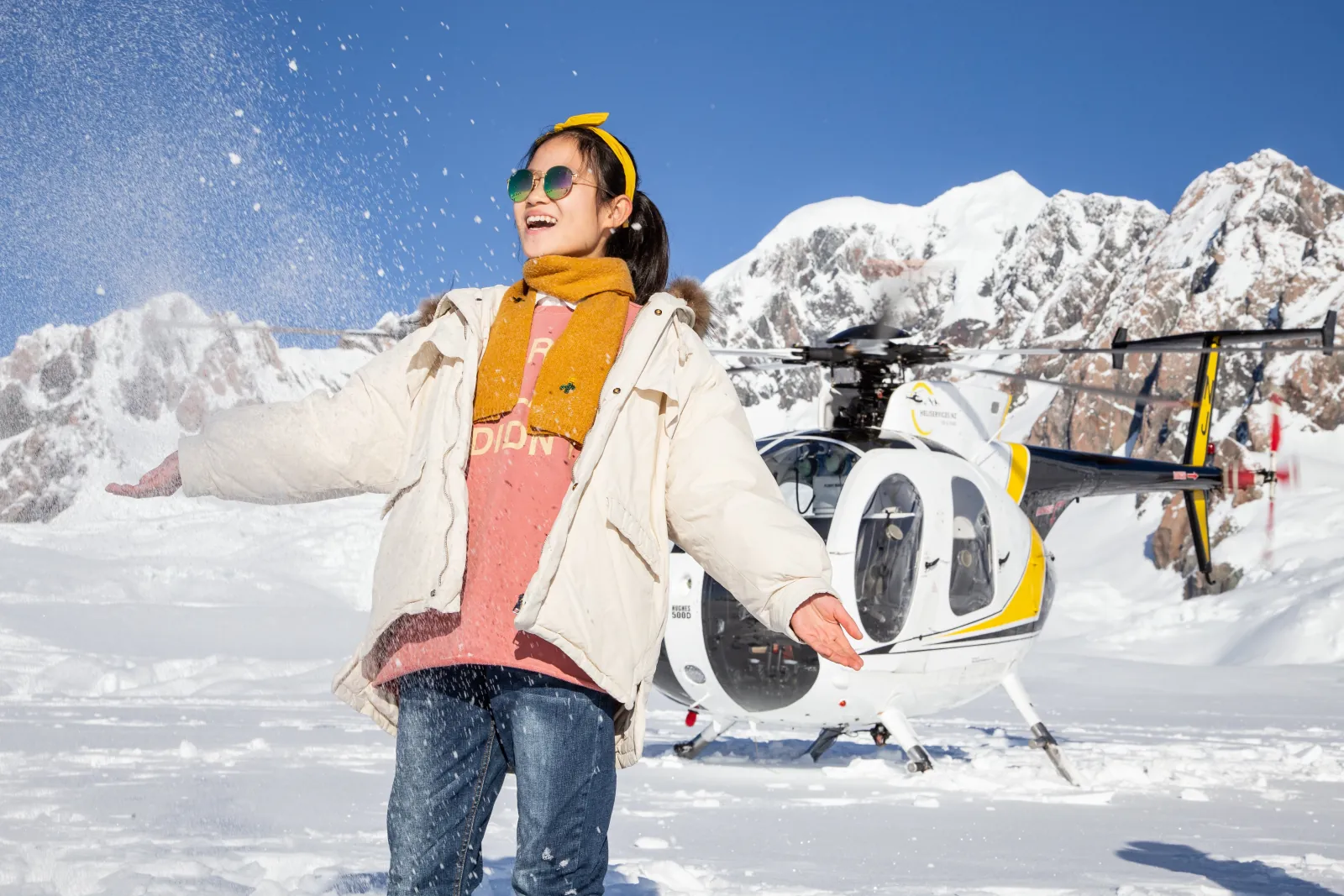 Girl in cream jacket with yellow scarf tossing snow beside a yellow and black helicopter