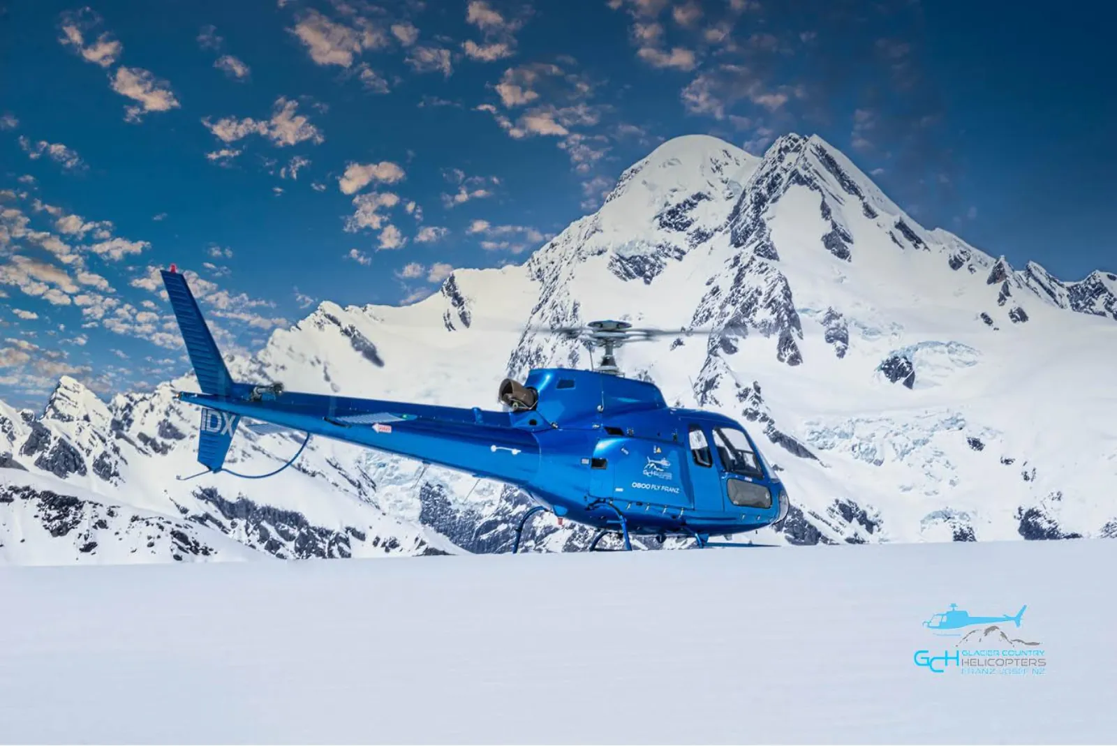 Blue helicopter parked on snow with a large snow-covered peak behind and scattered clouds