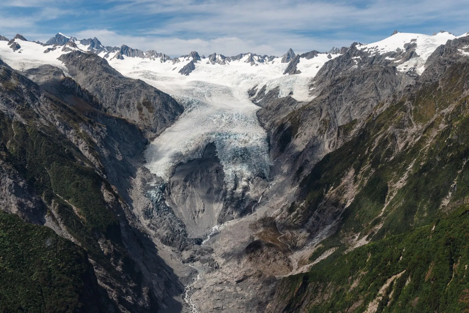 Aerial view of a glacier tongue descending a rocky valley between snowy peaks