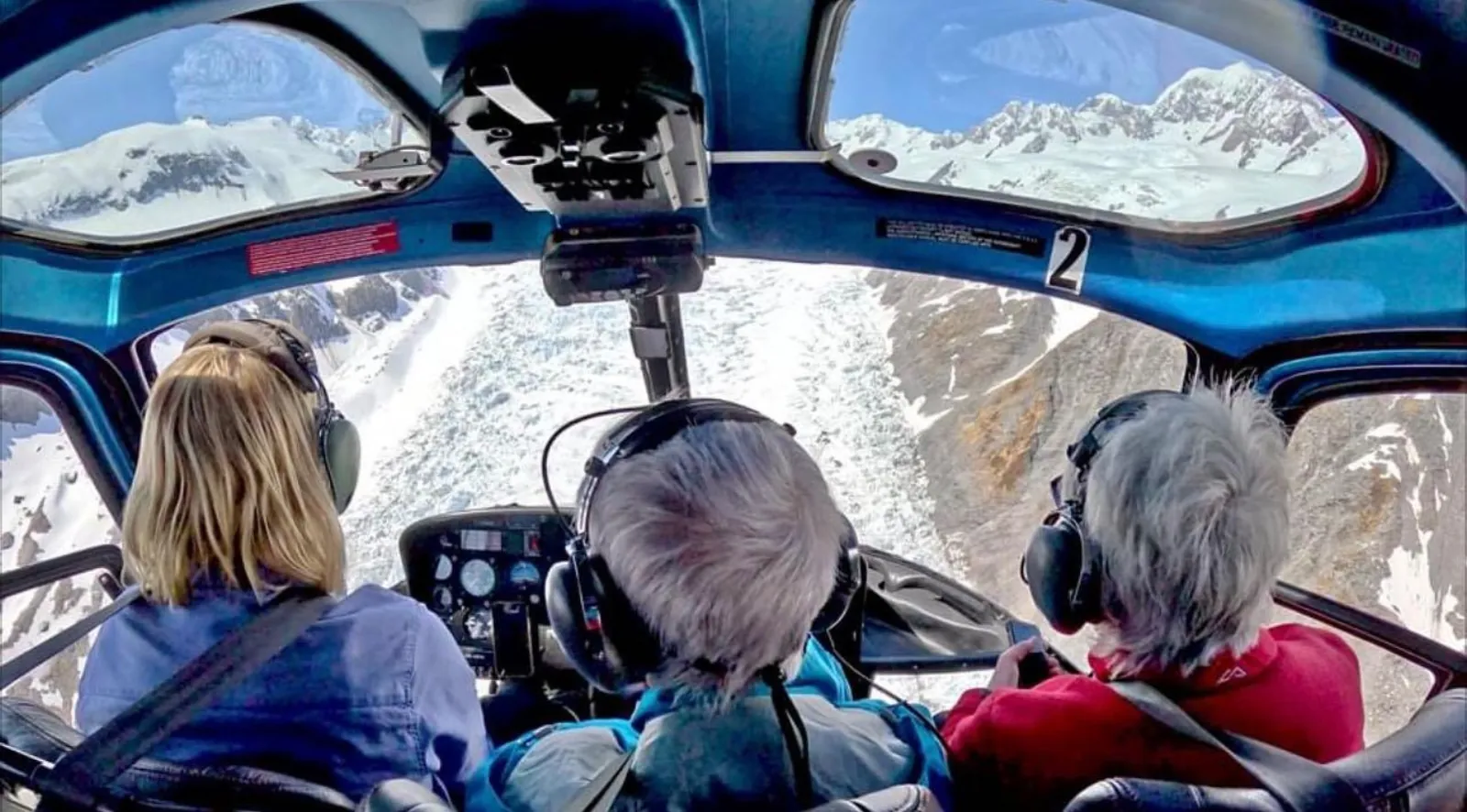 Passengers wearing headsets in a helicopter cockpit looking out at glacier ice and mountains