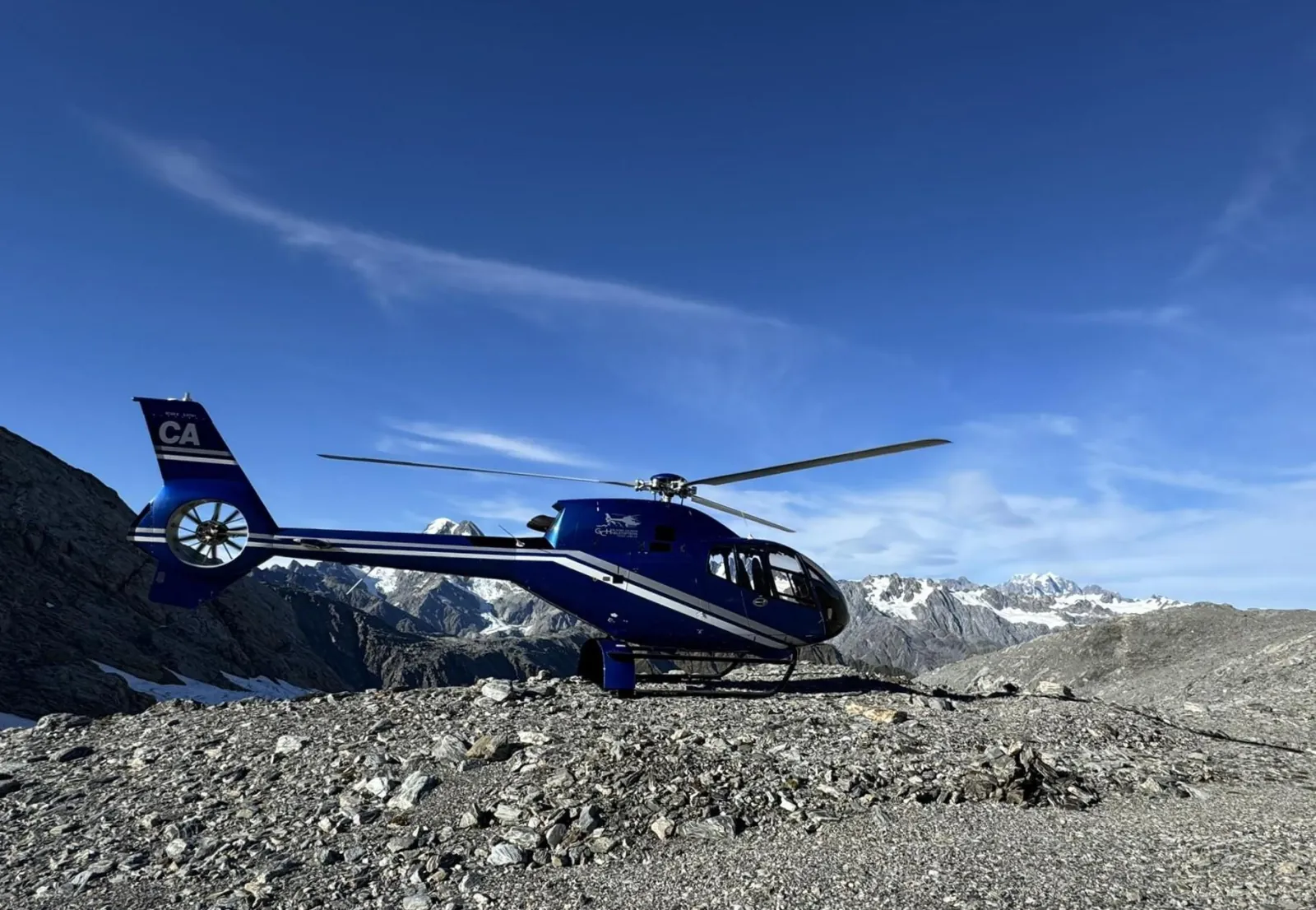 Dark blue helicopter landed on grey gravel beside a glacier with mountains behind
