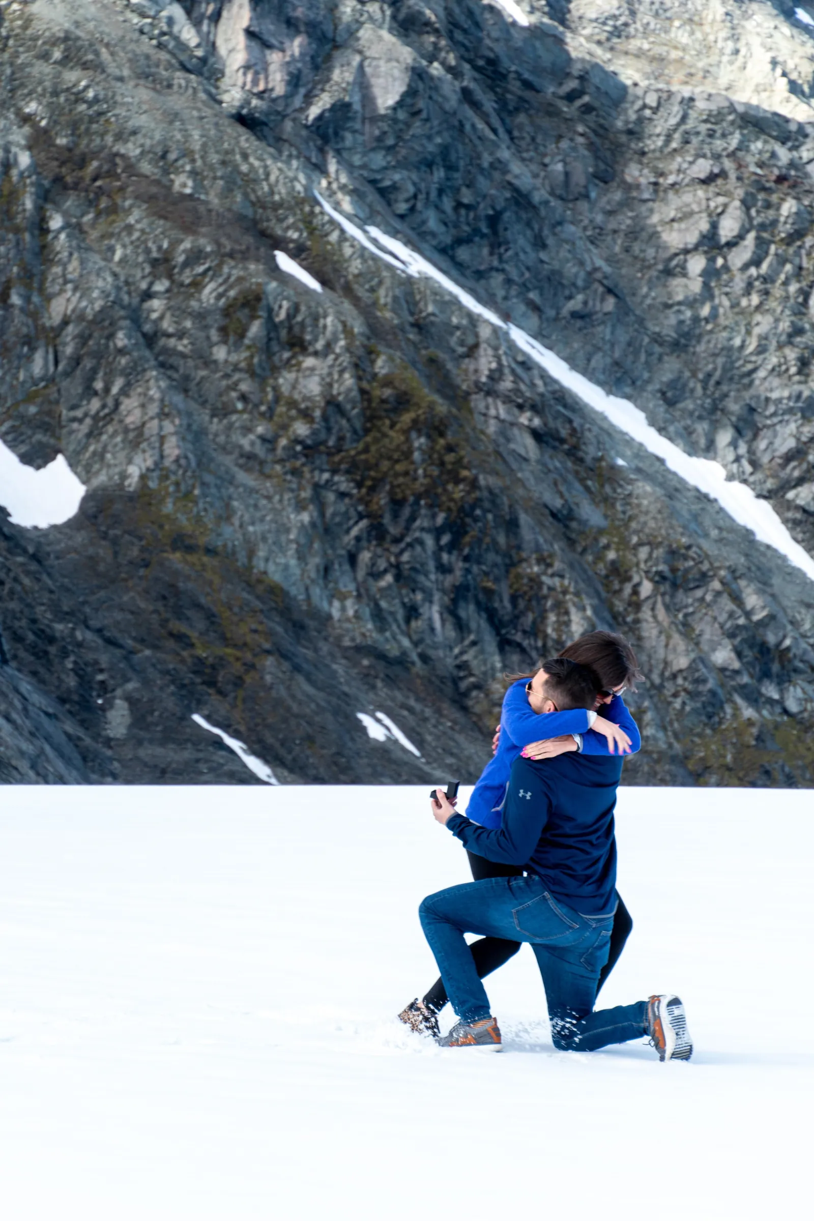 Man in blue jacket kneeling on snow in front of a rock face