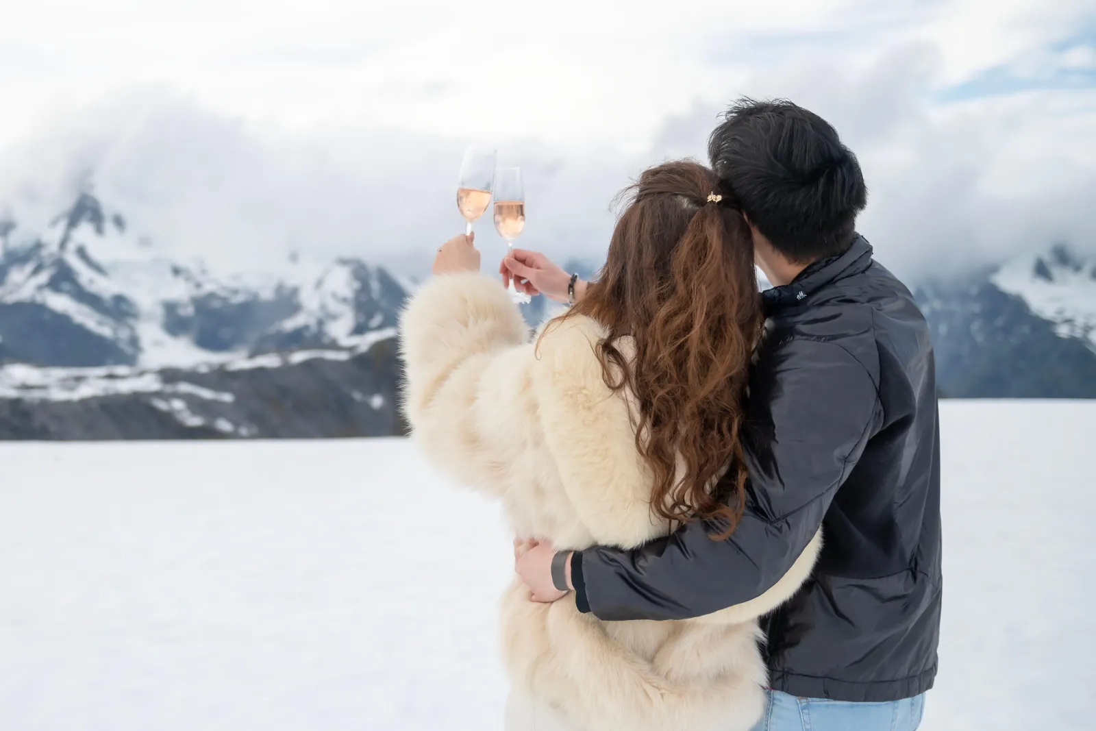 Couple holding up champagne glasses in an embrace on snow in front of cloudy peaks