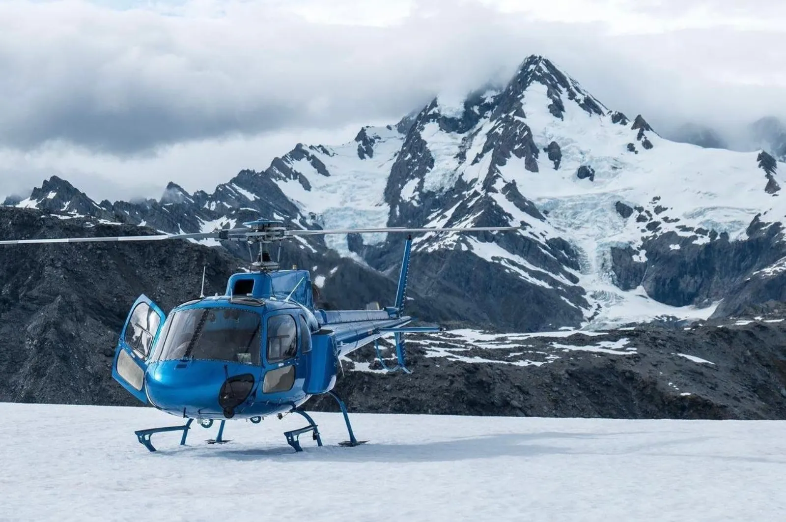 Blue helicopter on snow in front of a cloud-capped mountain with a hanging glacier