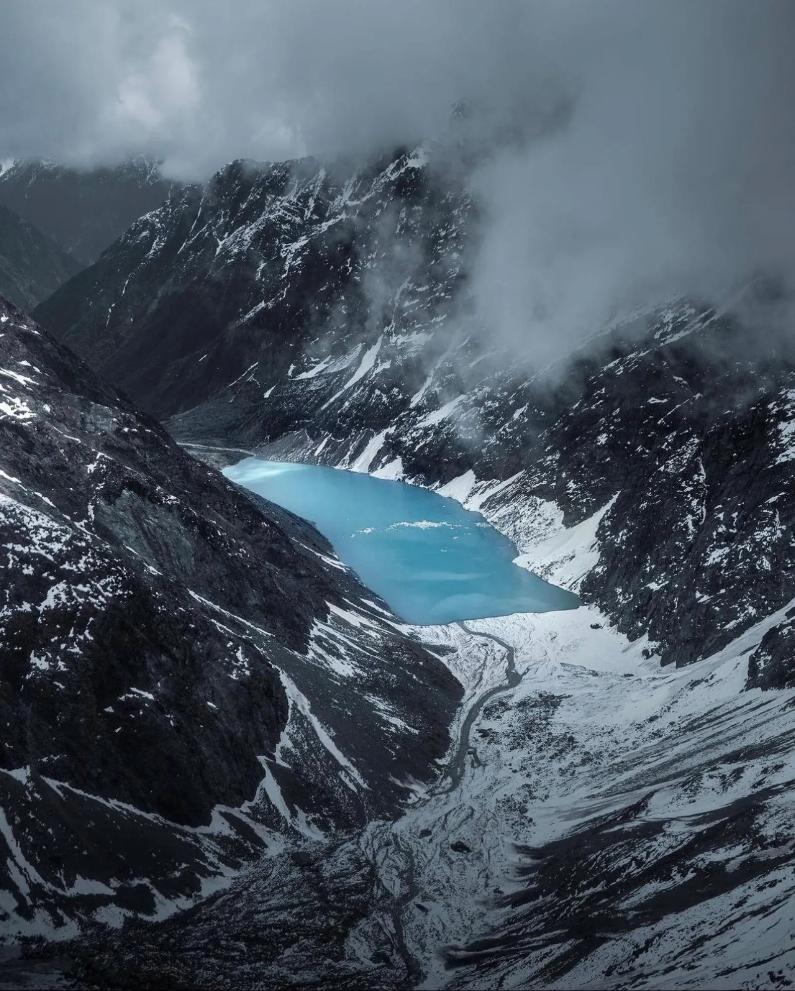 Aerial view of a turquoise glacial lake in a rocky valley beneath cloud