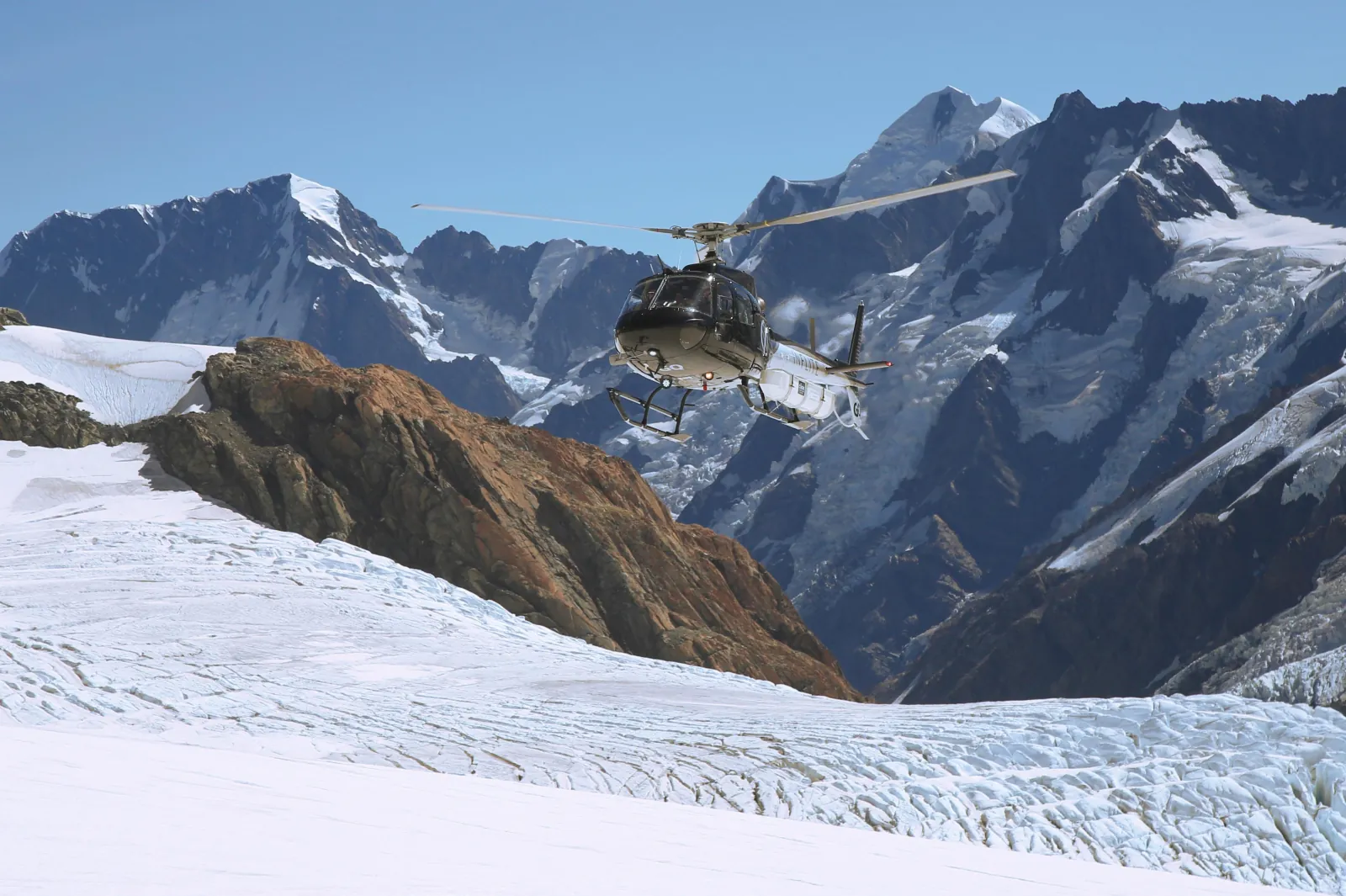Dark helicopter banking past a brown rocky outcrop and snowy ridges in strong light