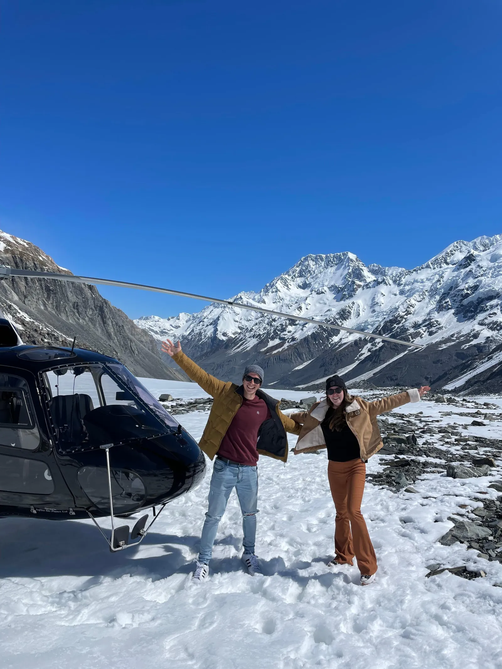 Two people with raised arms by a dark helicopter on snow with blue sky