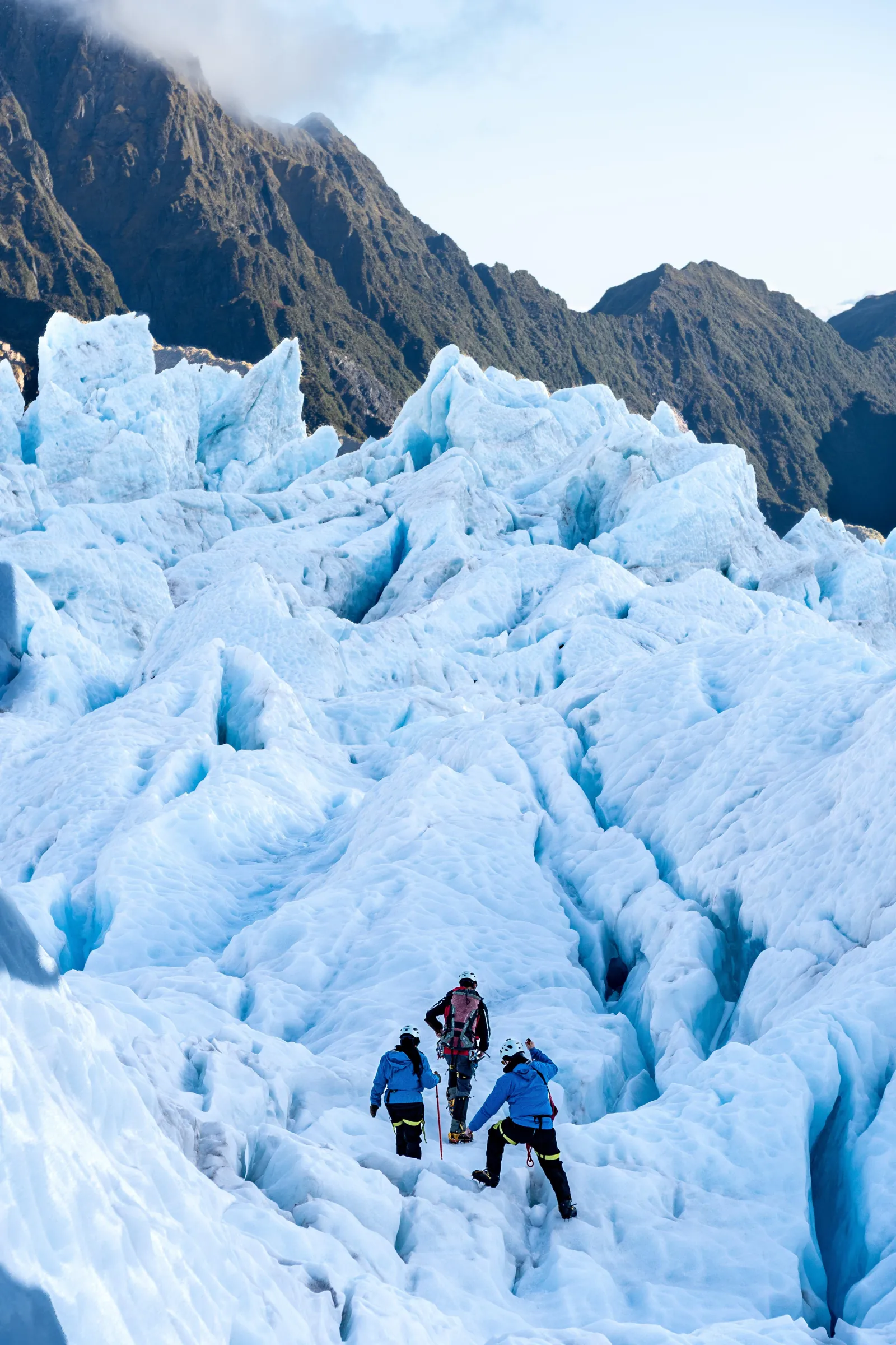 Two tiny hikers crossing a steep field of blue ice towers below dark rock